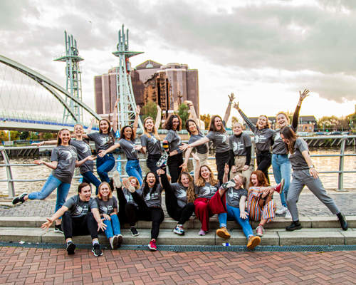 20 male and female dance ambassadors posing in front of Manchester bridge and skyline in the background. Wearing grey Dance ambassador tshirts.