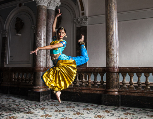 South Asian dancer wearing traditional blue and gold trousers and cropped top with bells on the ankles and a gold jewellery headpiece. Jumping in the air with point toe and one arm above the head and the other crossed over to the left. In front of the museum marble staircase.
