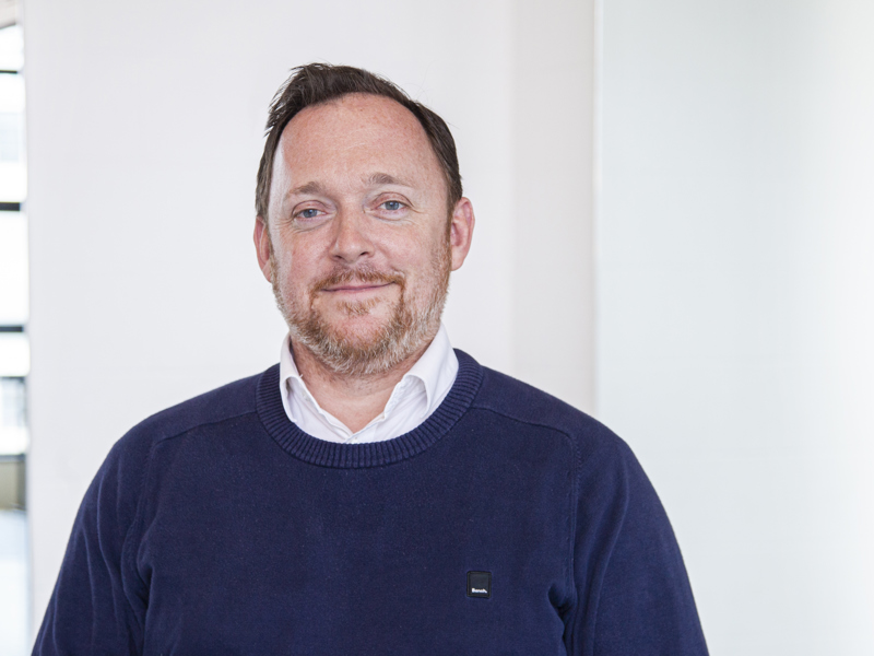 Headshot of Andrew Hurst. White male with light hair and beard smiling at camera, wearing blue jumper 