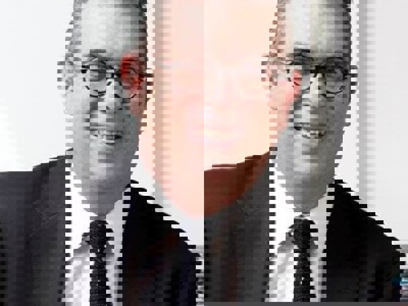 Headshot Anthony Bowne. White male smiling with circular glasses. Short grey hair, wearing a black suit, white shirt, and black tie.