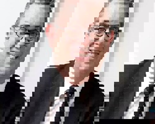 Headshot Anthony Bowne. White male smiling with circular glasses. Short grey hair, wearing a black suit, white shirt, and black tie.