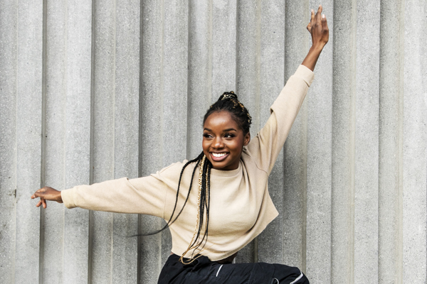 Global majority female with long braided hair standing on one leg with one arm above head and one arm to the side. Wearing black trousers and cream jumper. In front of grey wall