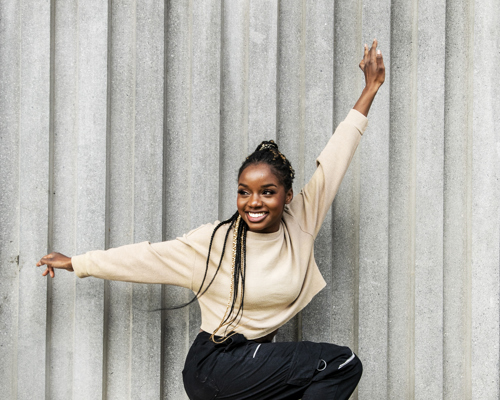 Global majority female with long braided hair standing on one leg with one arm above head and one arm to the side. Wearing black trousers and cream jumper. In front of grey wall