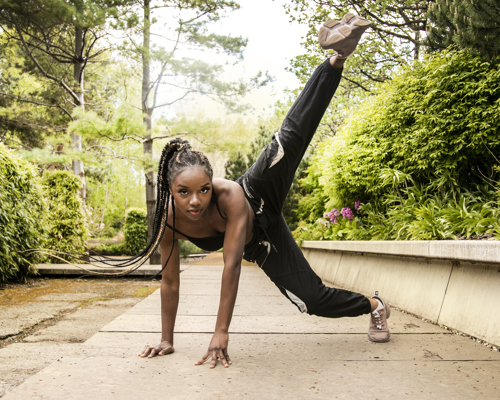 Global majority female hands on the floor kicking leg in the air with braided hair flicking to the left. Wearing black trousers and strap top. In a garden on a path