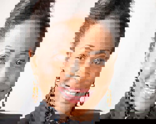 Headshot of Denise Nurse. Global majority female with tied back dark hair, smiling. Wearing large hoop earrings, black blazer and ruffled black top. 