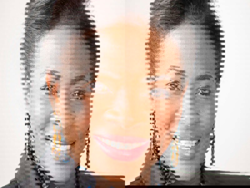 Headshot of Denise Nurse. Global majority female with tied back dark hair, smiling. Wearing large hoop earrings, black blazer and ruffled black top. 