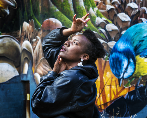 Global Majority female looking up to the top left of the image with hands around her face. Wearing a black leather jacket with short afro hair. In front of colourful graffiti wall