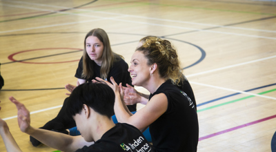 white female teacher with teenage student sitting on a sports hall floor with hands gesturing. All wearing black with linden dance logo on the t-shirts. 