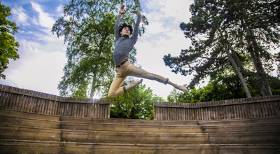 global majority male dancer jumping high with arms above head. Wearing grey long sleve tshirt and beige trousers.  In front of soft focus trees and blue sky.