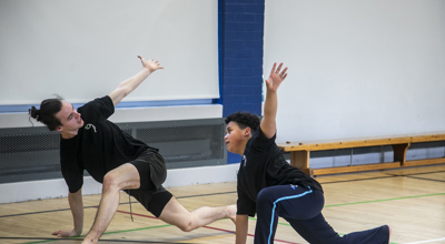 teenage male dance teacher lunging with one arm in the air teaching young boy dancer lunging with the mirrored arm in the air. In sports hall both wearing black PE kit