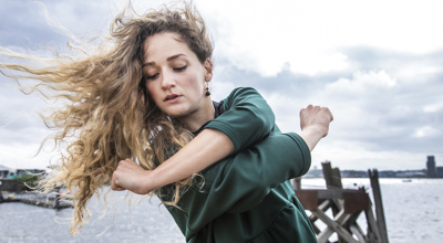 white female with arms crossed over body and long blonde curly hair flying in the wind to the left. In front of the sea and cloudy sky