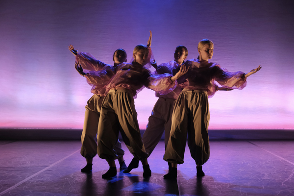 4 female dancers wearing baggy pink tops and cargo trousers all huddled together with arms stretched out leaning towards eachother. Stage lighting is pink and purple