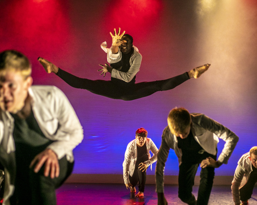 male dancers on U.dance stage wearing white shirts and black trousers. Four white young males crouched down one male black dancer jumpinging the box splits leaning forward with one hand stretched out. Lighting pink blue and red spotlights
