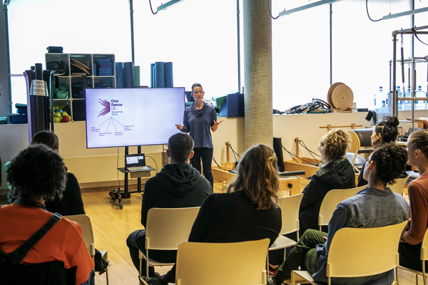 Hispanic female with tied up dark hair presenting with a screen behind her with One Dance UK logo on it. To a room of young people looking at her in an excersise classroom