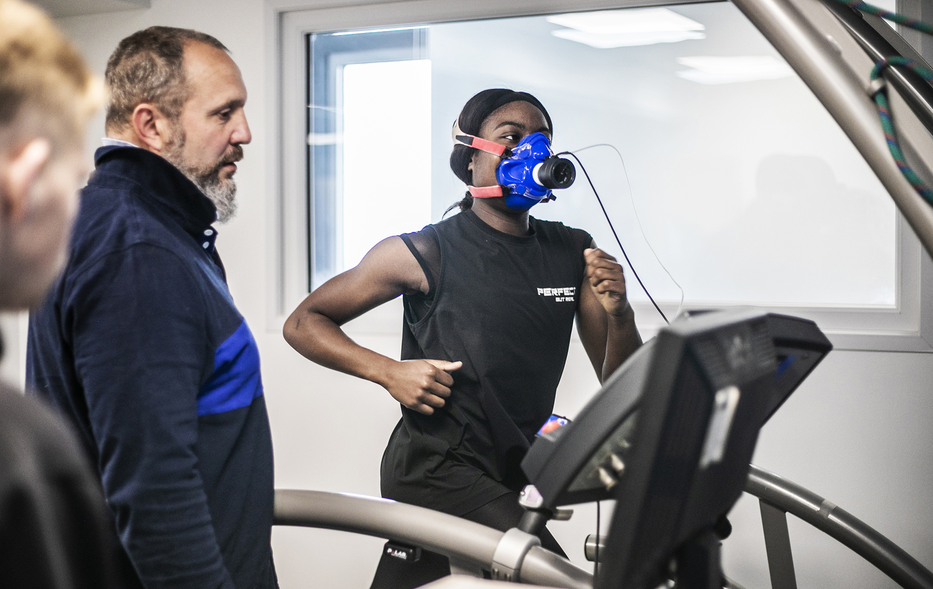 Global majority female dancer running on a treadmill wearing face-mask to measure cardiovascular fitness. White male physiologist looking over to observe stats on the machine. In a white laboratory setting with a window in the background