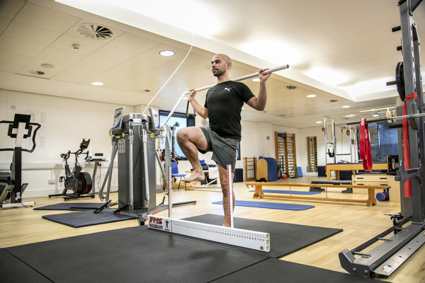 A bald male standing on one leg stepping over floor ruler, holding a pole behind his back. In a gym with lots of excersise equpiment in the background 
