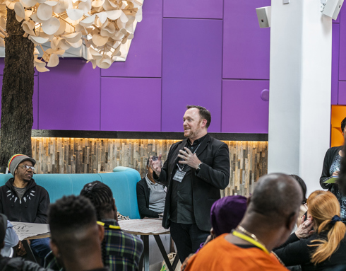 Centre of the image Andrew Hurst director of One Dance UK, white male standing in a colourful piurple room speaking to a group of people mixed genders, ages and race sitting and standing up around him. 