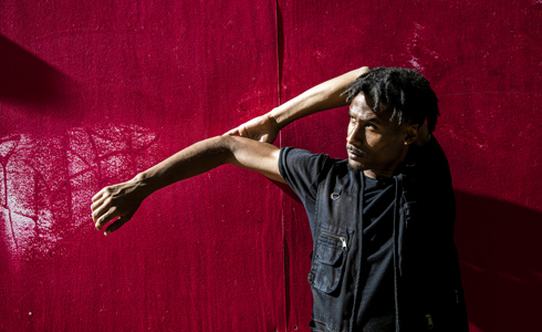 Global majority male dancer with short dreaded hair, one arm strecthed to the left and the other holding the elbow behind his head. Wearing all black standing in front of a red wall .