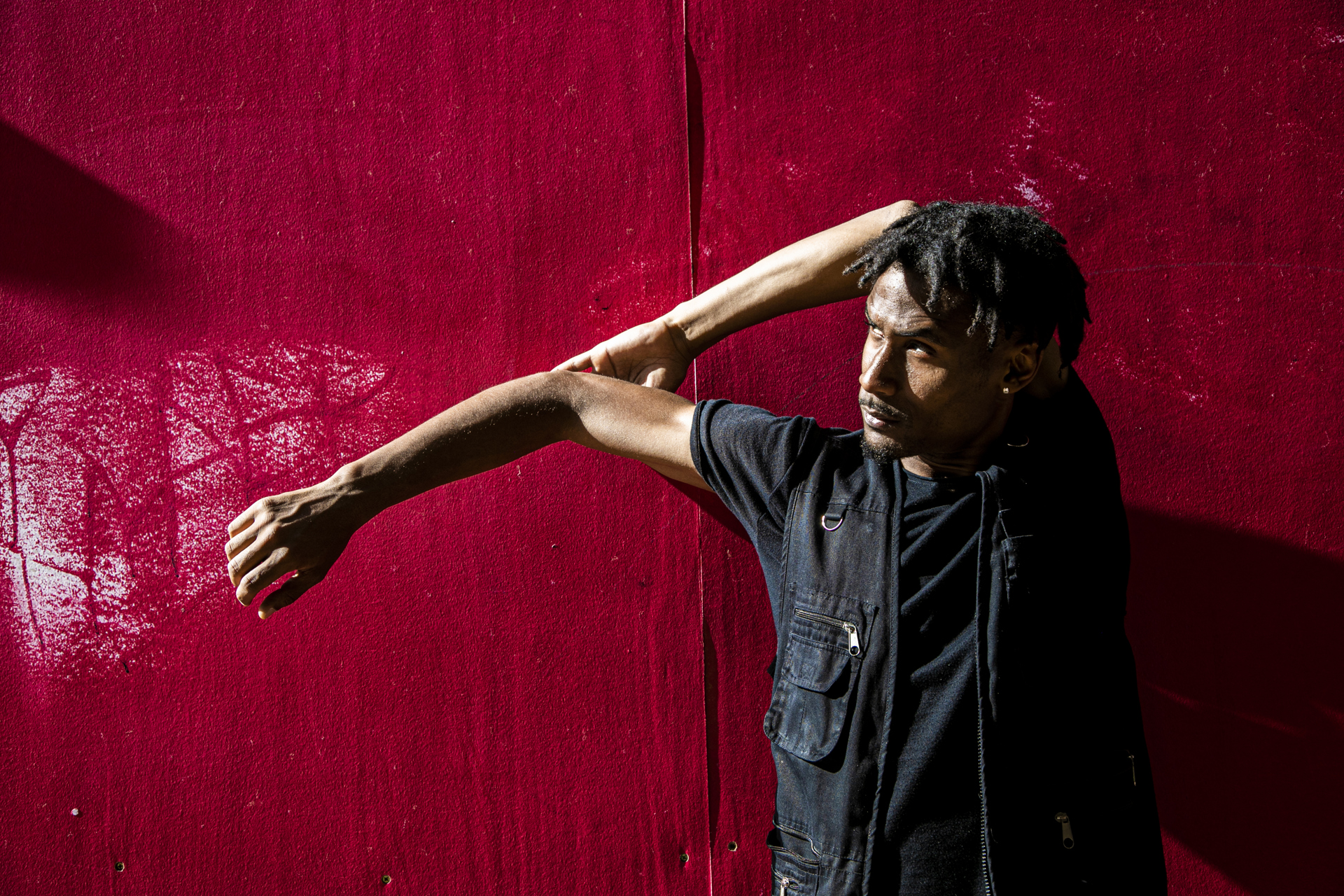 Global majority male dancer with short dreaded hair, one arm strecthed to the left and the other holding the elbow behind his head. Wearing all black standing in front of a red wall .