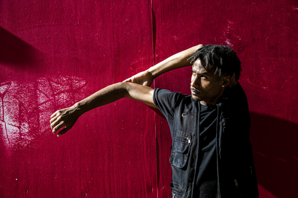 Global majority male dancer with short dreaded hair, one arm strecthed to the left and the other holding the elbow behind his head. Wearing all black standing in front of a red wall .