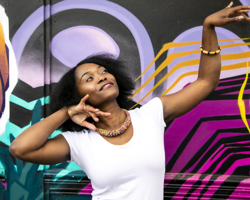 Global majority female dancer with afro, holding one arm out to the top right of the image with eyes looking at a limp wrist and the other hand next to her face. Wearing white top and colourful beaded necklace in fornt of coloured graffiti wall