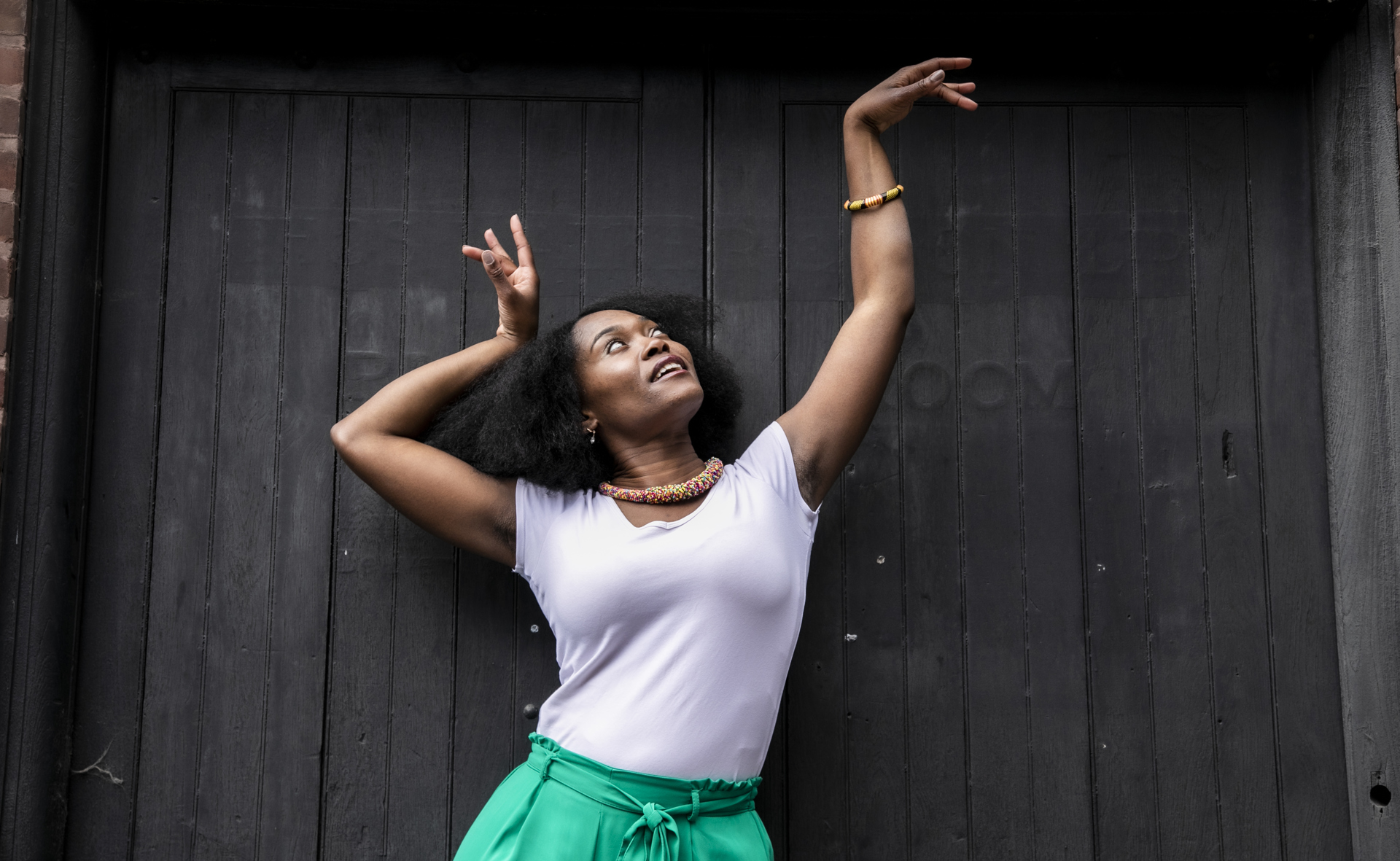 Global majority female dancer with afro, holding one arm out to the top right of the image with eyes looking at a limp wrist and the other hand next to her head. Wearing white top, green trousers and colourful beaded necklace in fornt of black wall