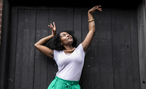 Global majority female dancer with afro, holding one arm out to the top right of the image with eyes looking at a limp wrist and the other hand next to her head. Wearing white top, green trousers and colourful beaded necklace in fornt of black wall