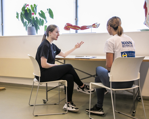 White female with brown tied back hair wearing black sitting at a table on white chair gesturing towards female dancer whos face is turned away from the camera sitting on white chair. In a room with large bright windows with a hanging skelaton and plant on window sill. 