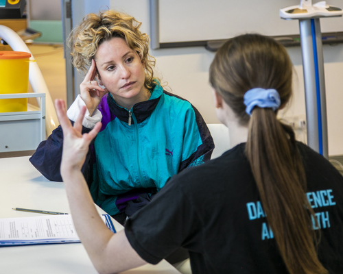 The back of white female with brown tied back hair wearing black sitting at a table gesturing towards white female dancer with curcly blonde hair and blue retro jacket leaning on one hand listening. with clipboard on the table and excersise equipment in the background in white room.