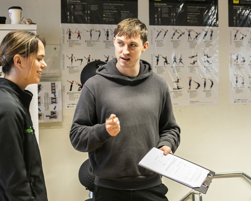 White male healthcare practitioner holding clipboard explainig something to white female in a healthcare environment with excersise posters in the background