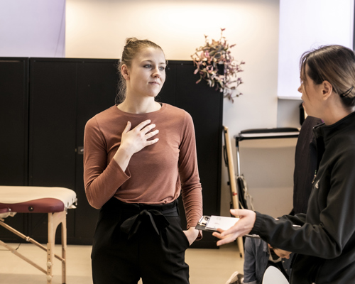 White female healthcare practitioner holding clipboard explainig something to white female dancer who is holding hand to heart in a healthcare environment with physio table in the background.