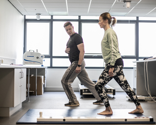 white male physio lunging forward holding his stomach, with white female dancer mirroring his movement. Both wearing excersise clothes in a bright healthcare labratory with large windows in the background.