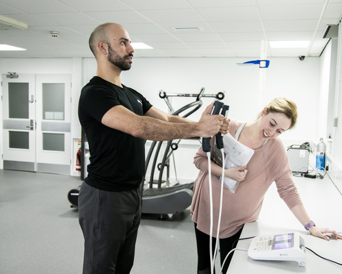 A male dancer is having his body composition measured by standing on a small scale with his arms outstretched in front of him. In either hand he is grasping a body-composition measurement device which are each connected to the scale with white cables. A white female screening assistant is leant over a white desk observing the monitor, and holding some papers. They are in a white and grey dance laboratory, with gym equipment in the background. 