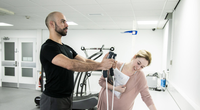 A male dancer is having his body composition measured by standing on a small scale with his arms outstretched in front of him. In either hand he is grasping a body-composition measurement device which are each connected to the scale with white cables. A white female screening assistant is leant over a white desk observing the monitor, and holding some papers. They are in a white and grey dance laboratory, with gym equipment in the background. 