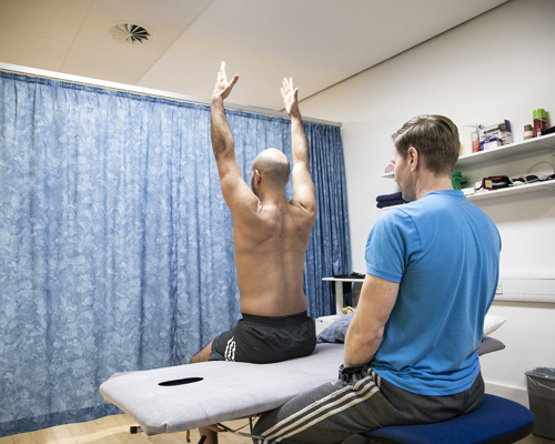 Male dancer sitting on physio bed with back to camera and arms up in the air, with male physiotherapist looking at dancer in an examination. With blue curtain in the background and a shelf of physio equipment. 
