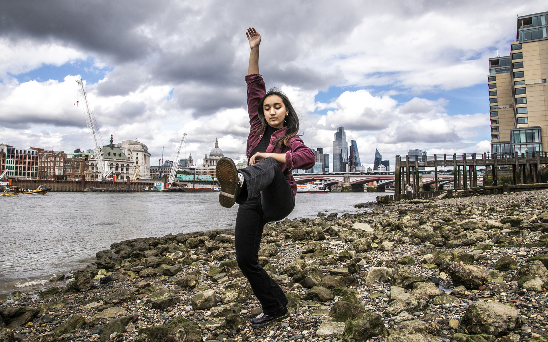 Female contemporary dancer with one leg risen and one arm above the head. Wearing black trousers and red open shirt standing with london beach skyline 