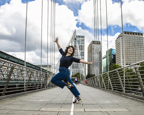 Young dancer with knee up one arm in the air one are to the side dancing on London Bridge. White female with short brown curly hair, wearing black shirt and blue jeans. 