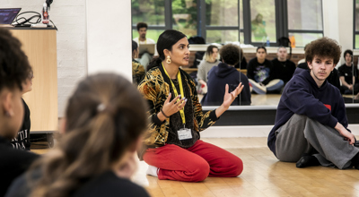 golbal majority dance teacher sitting on her knees speaking to a room full of dance students gesturing her hands, with mirror reflecting the students behind her. Wearing red trousers and colourful jacket. 