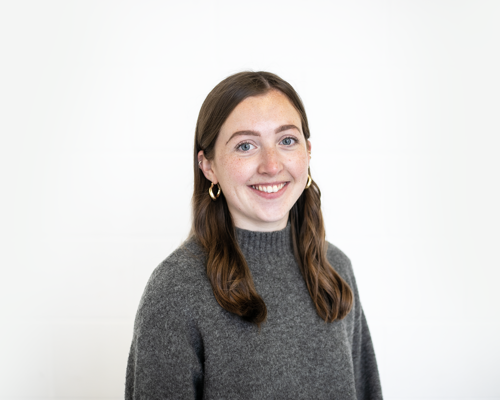 Headshot of Chloe Sprackling white female with brown hair smiling at the camera. Wearing gold hoop earrings and high neck grey jumper