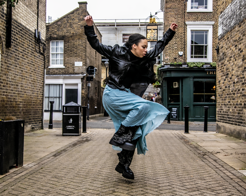 Global majority female dancer skipping down a london street side on with arms in the air . Wearing black leather jacket and blue skirt