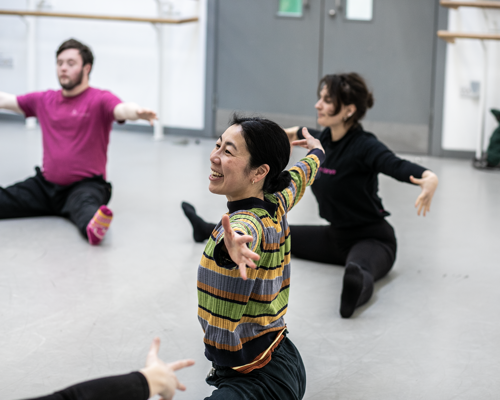 global majority female dancer teaching community group. Arm stretched open sitting with legs wise on the floor in the dance studio. 