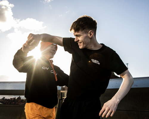 two young male dancers dancing together holding hands with lens flare inbetween them. wearing black and orange with one dancer in a beanie hat. infront of cloudy sky. 