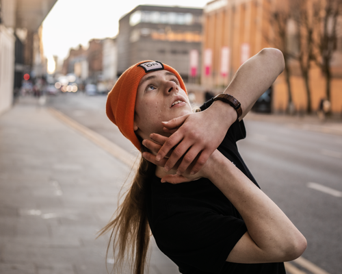 white male dancer with long blonde hair and orage beanie hat and black t shirt. On the street with hand around the face 