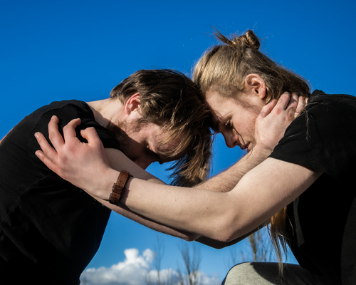 Two white male dancers holding eachothers forearms with heads together looking down with blue sky background.