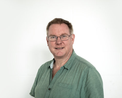 Headshot of Paul Hibbert. White light haired  male smiling at the camera wearing glasses and a green shirt